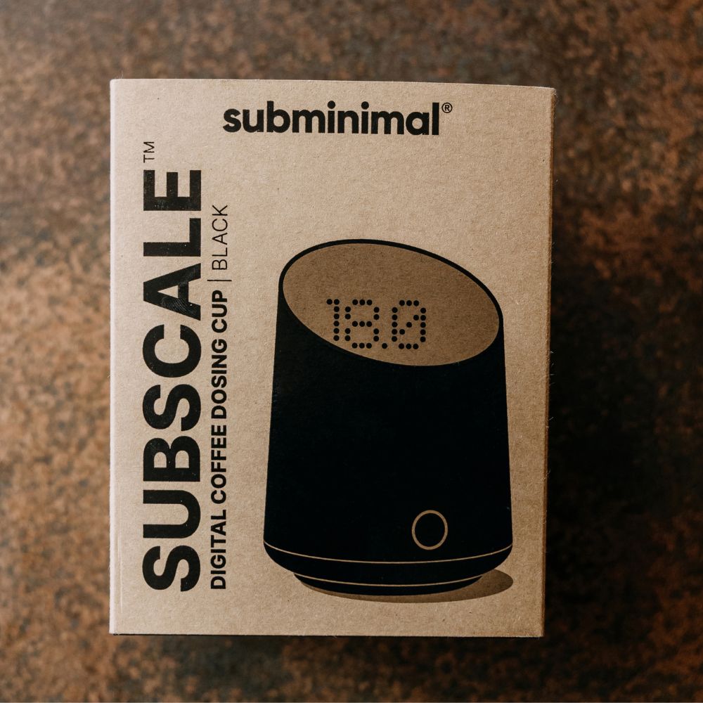 A box of subminimal Subscale digital coffee dosing cups in black, photographed from above on a dark textured surface. The packaging is kraft paper brown with black text and an illustration of the black cup showing a digital display.
