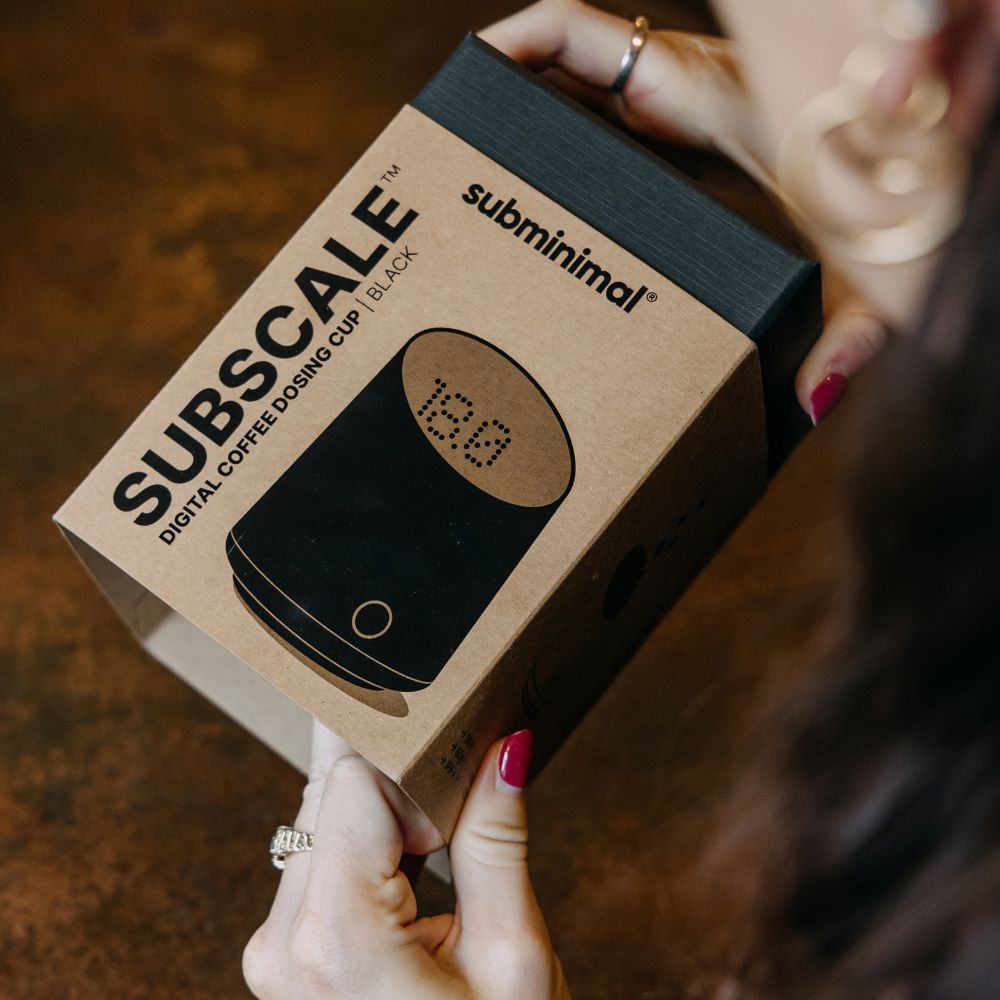 A person holds a brown cardboard box for the subminimal Subscale digital coffee dosing cup in black. The box features a graphic of the black cup with a digital display showing '12.